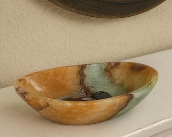 May include: Oval-shaped onyx bowl with a natural color gradient of tan, brown, and green. The bowl contains keys and a small black object. The bowl is on a white surface, with a dark-framed mirror in the background.