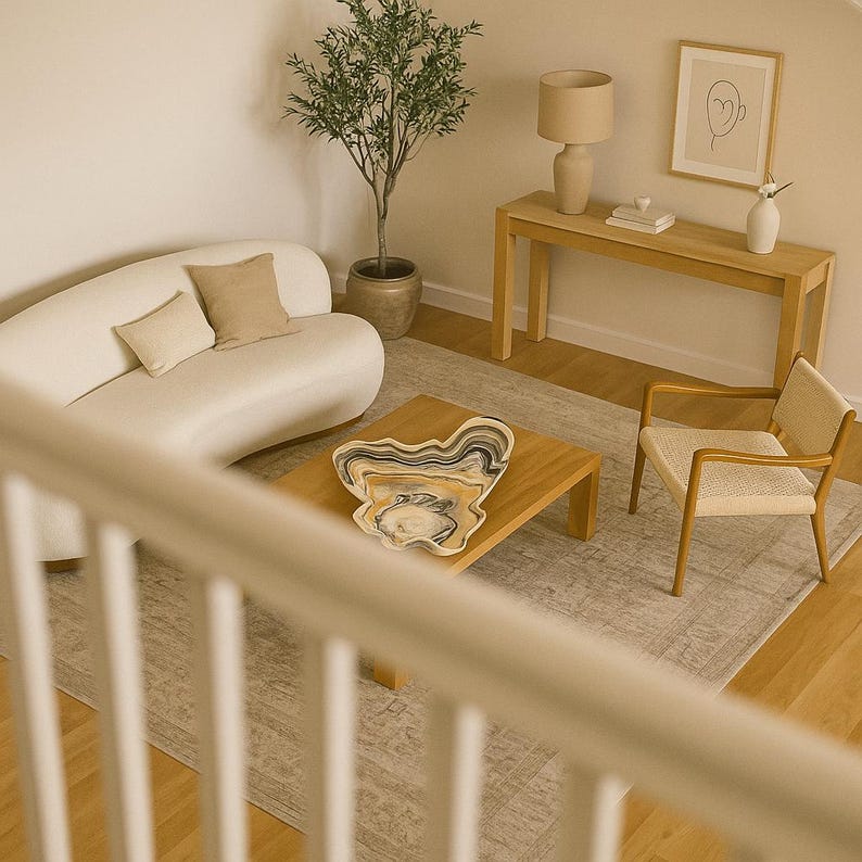 May include: A modern living room with a curved white sofa, wooden coffee table, and a decorative bowl. A beige rug, wooden side table, and a framed artwork complete the space. A small tree and a beige lamp add to the decor.