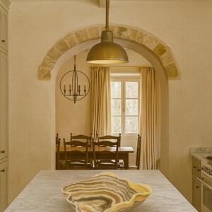 May include: A large, organic-shaped bowl made of banded stone with swirling layers of cream, brown, gray, and black. The bowl features a polished, wavy rim and sits centered on a white marble kitchen island countertop in a room with an arched doorway.