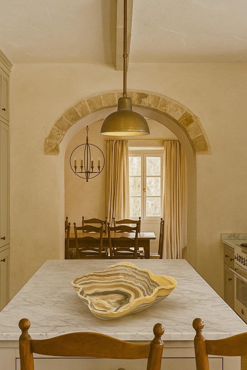 May include: A large, organic-shaped bowl made of banded stone with swirling layers of cream, brown, gray, and black. The bowl features a polished, wavy rim and sits centered on a white marble kitchen island countertop in a room with an arched doorway.