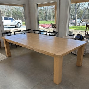 May include: A large, rectangular wooden dining table with a light oak finish. The table has thick, square legs and a smooth surface. The table is set in a room with large windows and several black chairs.