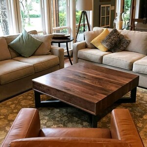 May include: A large, square, wooden coffee table with a dark walnut finish and a black metal frame. The table is in a living room setting with two couches and a rug.