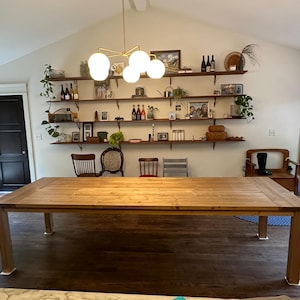 May include: A large wooden dining table with a natural wood finish. The table has a rectangular top and four legs. The table is in a dining room with a white wall and a set of shelves with various items on them.