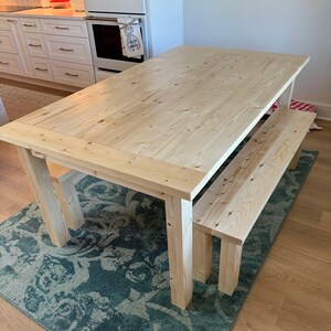 May include: A light wood dining table with a matching bench. The table has a simple design with a rectangular top and four legs. The bench is also rectangular with a simple design and two legs.