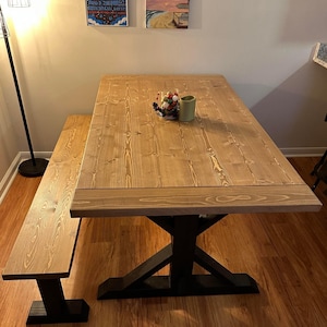 May include: A wooden dining table with a black X-shaped base and a matching bench. The table top is made of natural wood with a light brown finish. The table is set with a small arrangement of red and green berries and a candle.