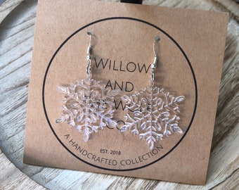 Puede incluir: Un par de pendientes de copo de nieve plateados con un acabado transparente y claro. Los pendientes están en una tarjeta que dice "Willow and The Woods Co. Est. 2018 A Handcrafted Collection."