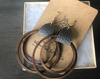 Puede incluir: Un par de pendientes de aro de madera y cuero gris. Los pendientes están hechos de tres aros de madera concéntricos y tienen una forma de lágrima de cuero gris que cuelga del aro superior. Los pendientes están empaquetados en una pequeña caja con un inserto de papel marrón que dice "Wow And The Wow Co."