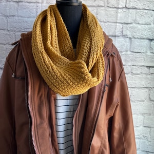 May include: A mustard yellow crocheted infinity scarf on a brown leather jacket with a white striped shirt underneath.