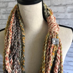 Puede incluir: Una bufanda infinita multicolor hecha de hilo de ganchillo en tonos de marr&oacute;n, beige, naranja y verde. La bufanda se muestra en un maniqu&iacute; y tiene una peque&ntilde;a etiqueta de cuero con el texto "*K*" en ella.