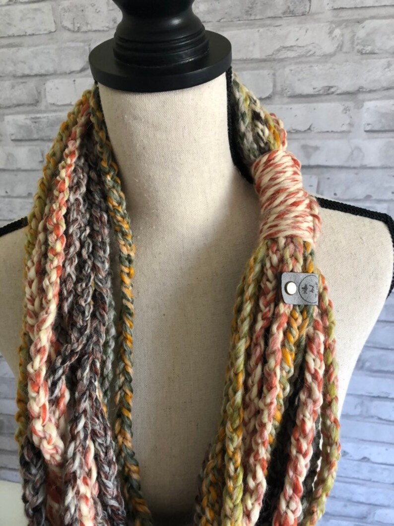 Puede incluir: Una bufanda infinita multicolor hecha de hilo de ganchillo en tonos de marr&oacute;n, beige, naranja y verde. La bufanda se muestra en un maniqu&iacute; y tiene una peque&ntilde;a etiqueta de cuero con el texto "*K*" en ella.
