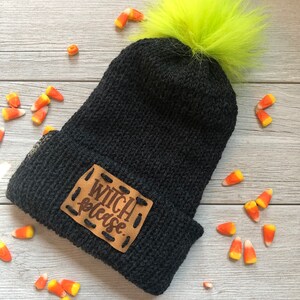 May include: Black knit beanie with a faux fur pom pom in bright green. The beanie has a brown leather patch with the text "Witch Please." sewn on.