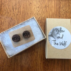 Puede incluir: Un par de pendientes de madera con la palabra "nope" grabada en cada pendiente. Los pendientes est&aacute;n empaquetados en una peque&ntilde;a caja marr&oacute;n con una etiqueta blanca que dice "Willow and The Wolf".
