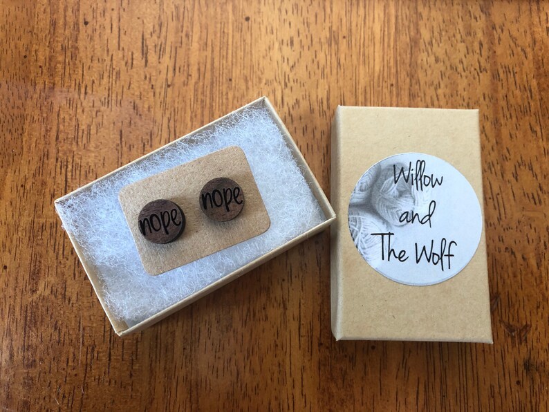 Puede incluir: Un par de pendientes de madera con la palabra "nope" grabada en cada pendiente. Los pendientes est&aacute;n empaquetados en una peque&ntilde;a caja marr&oacute;n con una etiqueta blanca que dice "Willow and The Wolf".