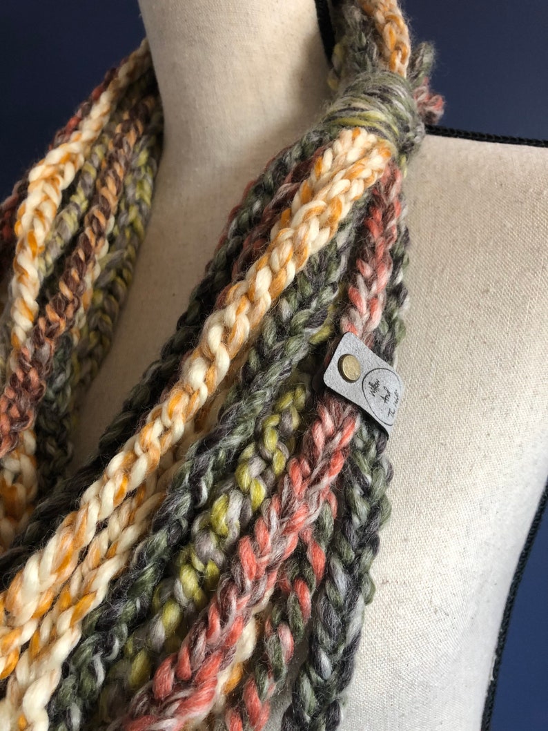 Puede incluir: Una bufanda infinita de crochet multicolor con una etiqueta de cuero marr&oacute;n que dice "Made with Love" en letras blancas.