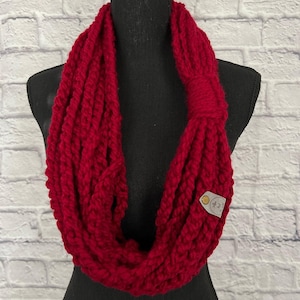 May include: A red infinity scarf made of chunky yarn. The scarf has a simple, textured design and is finished with a small leather tag.