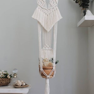 Puede incluir: Un macetero de macramé blanco con una barra de madera y una maceta marrón. El macetero está colgado de un gancho en una pared blanca. La maceta está llena de flores secas y vegetación.