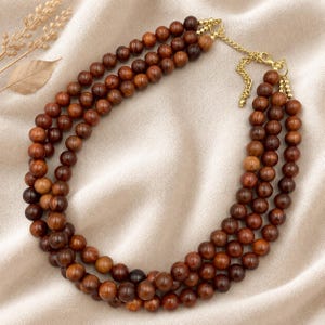 May include: A multi-strand necklace featuring three rows of polished brown wooden beads. The necklace is secured with a gold-toned metal clasp and includes an adjustable chain extension, providing a layered look with varying shades of brown.