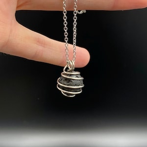 Könnte beinhalten: Eine silberfarbene Halskette mit einem dunkelgrauen, kugelförmigen Anhänger, der in einem Drahtkäfig eingeschlossen ist. Der Anhänger scheint ein poröser Stein zu sein, der an einer zarten Kette hängt. Der Hintergrund ist dunkel und einfarbig.