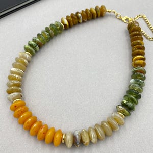 May include: A beaded necklace featuring a gradient of disc-shaped beads in shades of amber, olive green, and cream. The necklace includes small gold-toned metal spacers between some beads and finishes with a gold-toned metal chain and lobster clasp.