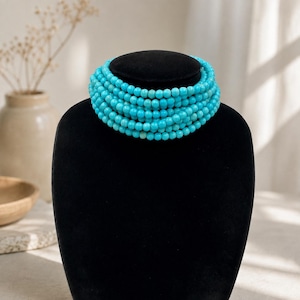 May include: A multi-strand necklace composed of numerous small, round, bright turquoise blue beads. The beads are uniform in size and shape, arranged in several tight, concentric circular layers that create a thick, layered appearance around the neck.