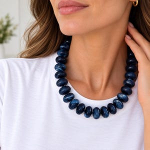May include: A necklace featuring a single strand of smooth, round, dark blue beads with subtle white marbled patterns. The beads are uniform in size and shape, creating a continuous circular design that rests around the neck against a white shirt.