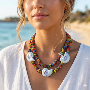 May include: A layered necklace with rainbow-colored beads and three seashell pendants. The necklace is worn against a white top, with a beach scene in the background.