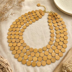 May include: A layered necklace featuring multiple strands of light brown wooden discs. The necklace has a gold-colored chain and clasp. The design is a statement piece, perfect for adding a natural touch to any outfit.