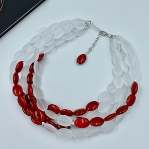 May include: A three-strand necklace featuring white and red oval beads. The necklace has a silver-toned chain and clasp. The beads are arranged in a layered design, with the red beads interspersed among the white ones.