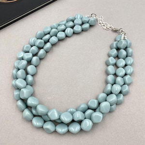 May include: A three-strand necklace featuring light blue, rounded beads. The beads are strung together with a silver-colored chain and clasp. The necklace is displayed on a neutral background.