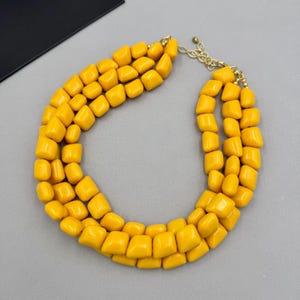 May include: A multi-strand necklace featuring three rows of smooth, chunky, mustard yellow beads with a glossy finish. The necklace is connected by a gold-toned metal chain link clasp, providing an adjustable length for the wearer.