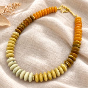 May include: A beaded necklace featuring a gradient of disc-shaped beads in shades of cream, yellow, ochre, and orange. The necklace includes small gold-toned metal spacers between some beads and a gold-toned metal chain extender with a lobster clasp.