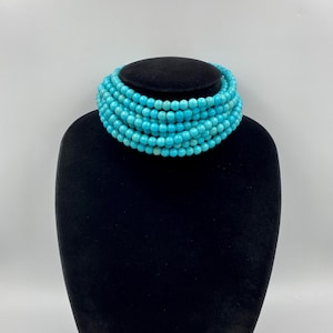 May include: A multi-strand turquoise bead necklace displayed on a black mannequin. The necklace is made of small, round, light blue beads. The necklace is wrapped around the mannequin's neck, showcasing its layered design.