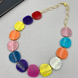 May include: A colorful necklace featuring large, irregularly shaped beads in a variety of colors, including red, purple, yellow, orange, pink, and blue. The beads are connected by a gold-colored chain.