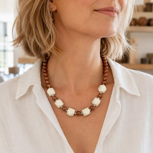 May include: A necklace featuring alternating cream-colored geometric beads and small, round, dark brown wooden beads. The necklace is worn around the neck and is set against a white shirt.