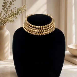 Puede incluir: Un collar tipo gargantilla de múltiples hilos con cinco filas de cuentas esféricas doradas y pulidas. Las cuentas son de tamaño uniforme y están dispuestas en una formación apretada y superpuesta, creando una banda circular continua.