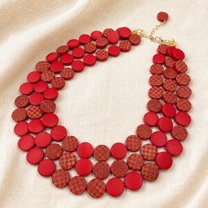 May include: A layered necklace featuring three strands of red, circular beads. Some beads have a checkered pattern in a complementary color. The necklace has a gold-colored chain and clasp.