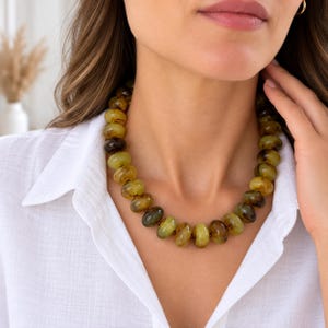 May include: A necklace featuring a series of smooth, rounded, translucent beads in varying shades of olive green, yellow, and dark brown. The beads are strung together in a single strand, creating a circular shape that rests around the neck.