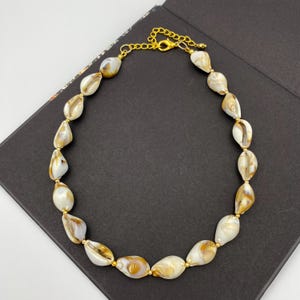 May include: A necklace featuring a strand of natural seashells in shades of white and brown, strung together with gold-colored metal beads and a chain clasp. The necklace is displayed on a dark surface.