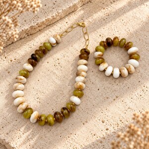 Puede incluir: Un conjunto de collar y pulsera con cuentas lisas, redondas y en forma de disco en tonos verde oliva, marrón oscuro y crema. El collar incluye una sección de cadena de metal dorado, mientras que la pulsera es un bucle continuo de cuentas.