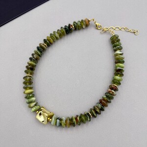 Puede incluir: Un collar con una hilera de pequeñas cuentas en forma de disco de color verde y marrón jaspeado, intercaladas con diminutos espaciadores dorados. Un dije de metal dorado más grande e irregular se encuentra en un lado del collar.