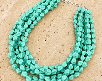 May include: A multi-strand statement necklace featuring numerous faceted, turquoise-colored beads with a matte finish. The necklace is secured with a silver-toned metal chain and a lobster claw clasp, creating a layered, voluminous appearance.