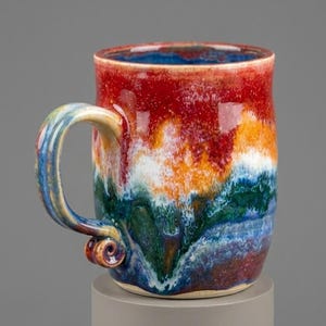 May include: A handmade ceramic mug with a colourful, layered design. The mug features bands of red, orange, white, green, and blue, with a blue interior and a curved handle. The mug is approximately 10 cm tall.