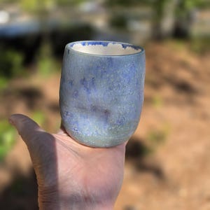 Puede incluir: Un vaso de cer&aacute;mica hecho a mano con un esmalte moteado azul y verde. La taza tiene una forma redondeada y un borde interior blanco. El vaso se sostiene en una mano sobre un fondo borroso al aire libre.