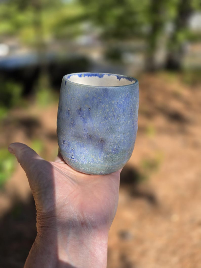 Puede incluir: Un vaso de cer&aacute;mica hecho a mano con un esmalte moteado azul y verde. La taza tiene una forma redondeada y un borde interior blanco. El vaso se sostiene en una mano sobre un fondo borroso al aire libre.