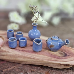 May include: Miniature blue ceramic tea set with gold trim, including a teapot, creamer, six cups, and a small vase with dried flowers. The set is displayed on a wooden tray, perfect for a dollhouse or decorative display.