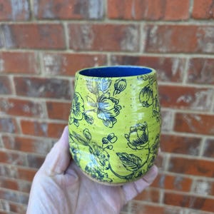 Puede incluir: Un vaso de cerámica hecho a mano con un vibrante esmalte amarillo-verdoso. El vaso está adornado con un patrón floral negro y tiene un interior azul. El vaso se sostiene en una mano contra un fondo de pared de ladrillo.