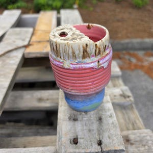 Puede incluir: Una taza de cerámica hecha a mano con un diseño único. La taza presenta una combinación de colores rojo, rosa, azul y verde. El borde superior es de color beige claro con goteos marrones. La taza tiene un agujero en la parte superior y una textura acanalada.