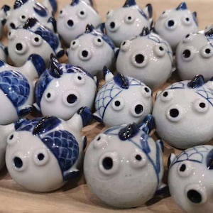 May include: A collection of ceramic fish figurines. Each figurine is white with blue accents, featuring detailed scales and fins. The fish have large, dark eye holes and are arranged closely together.