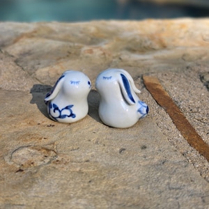 May include: Two small white ceramic rabbit figurines featuring hand-painted blue floral and vine patterns. Each rabbit has closed eyes and long ears, with one showing a detailed scroll design on its side and the other displaying blue ear accents.