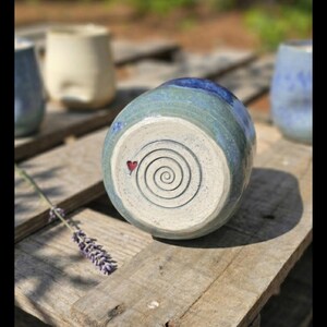 Puede incluir: Un jarr&oacute;n de cer&aacute;mica hecho a mano con un esmalte azul y verde, con un dise&ntilde;o en espiral y un peque&ntilde;o coraz&oacute;n rojo en la base. El jarr&oacute;n se exhibe sobre una superficie de madera r&uacute;stica, con otras piezas de cer&aacute;mica al fondo.